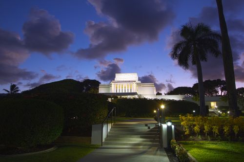 A set of stairs leading up to the Laie Hawaii Temple, which is seen in the distance atop a hill in front of a purple night sky.