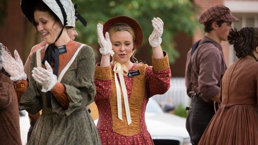 Young female missionaries walking on the street with their hands in a clapping motion dressed in pioneer era clothing.