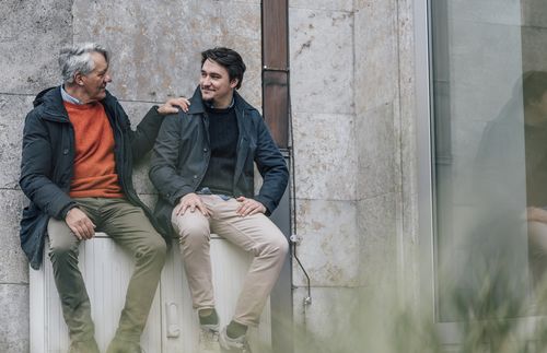 two men seated by a building talking to each other