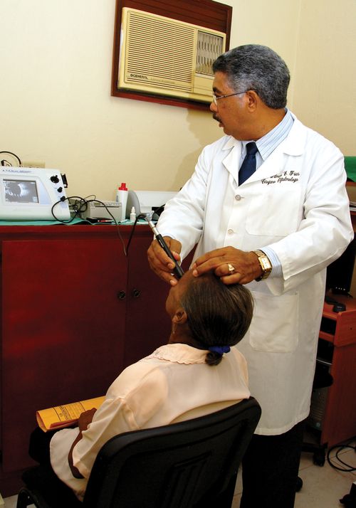 A male doctor in the Dominican Republic, wearing a long white coat, using a long metal device to check the eyesight of a woman sitting down.