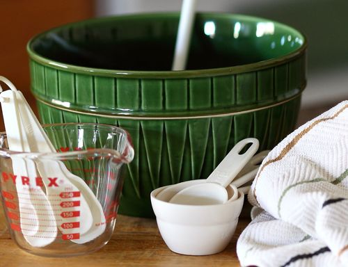 A table setting with a large green mixing bowl, measuring cups, and measuring spoons next to a dish towel.