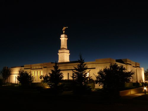 A night view of the Monticello Utah Temple lit up, with silhouetted trees in front.