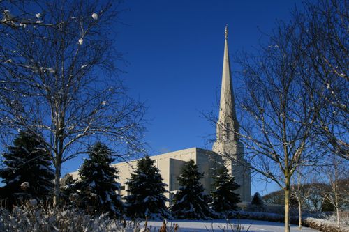 A side view of the Preston England Temple, seen between bare winter trees, over a snow-covered lawn, with a deep blue sky overhead.