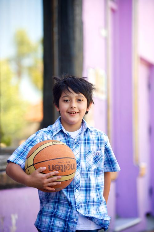 A young boy in a blue and white plaid shirt stands outside holding a basketball in one arm.