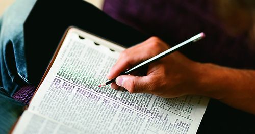 Hands of young man holding red marking pencil, while reading and studying open scriptures on lap. (horiz)