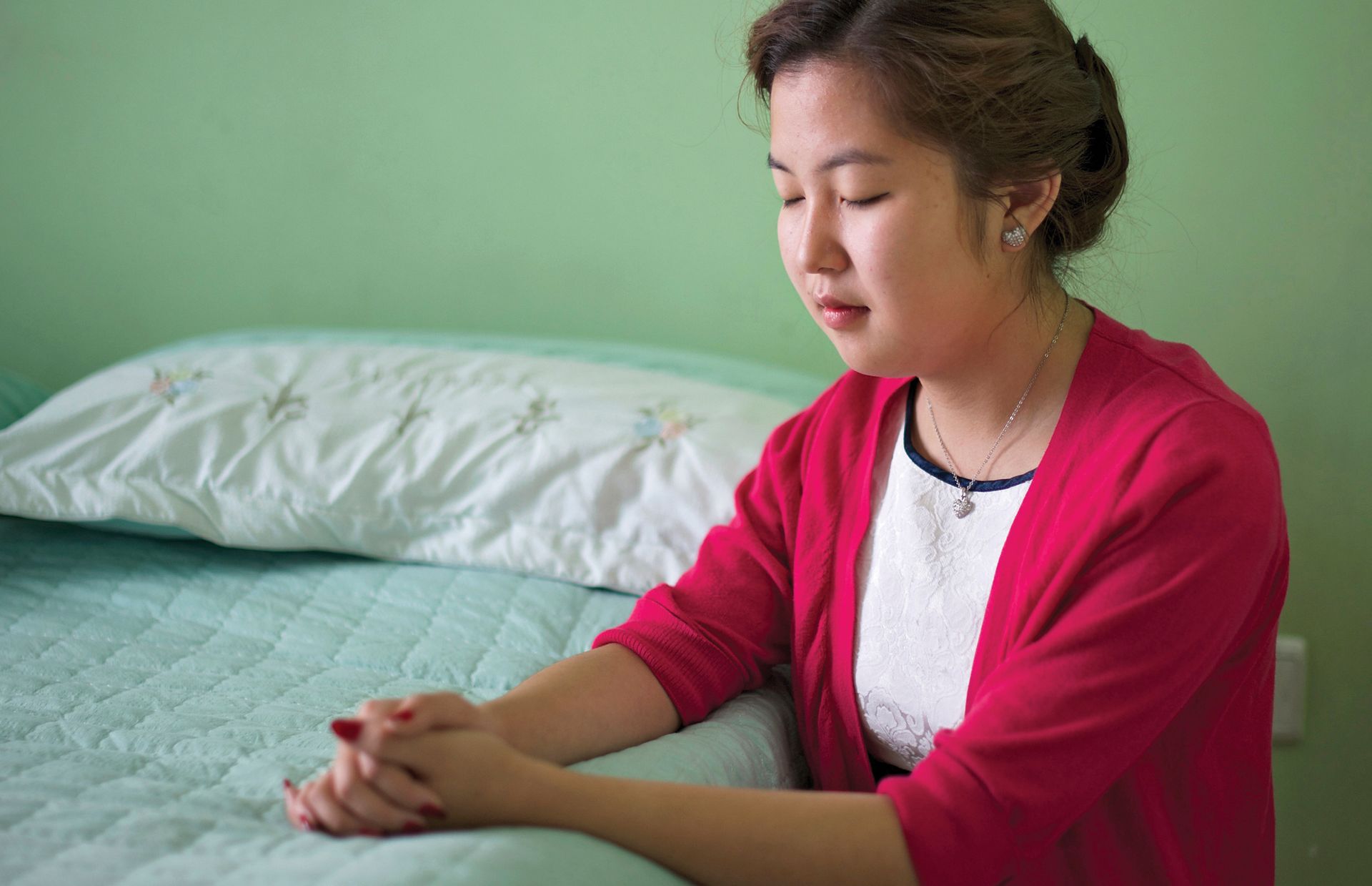 A young woman from Mongolia kneels by her bed, closes her eyes, and prays.