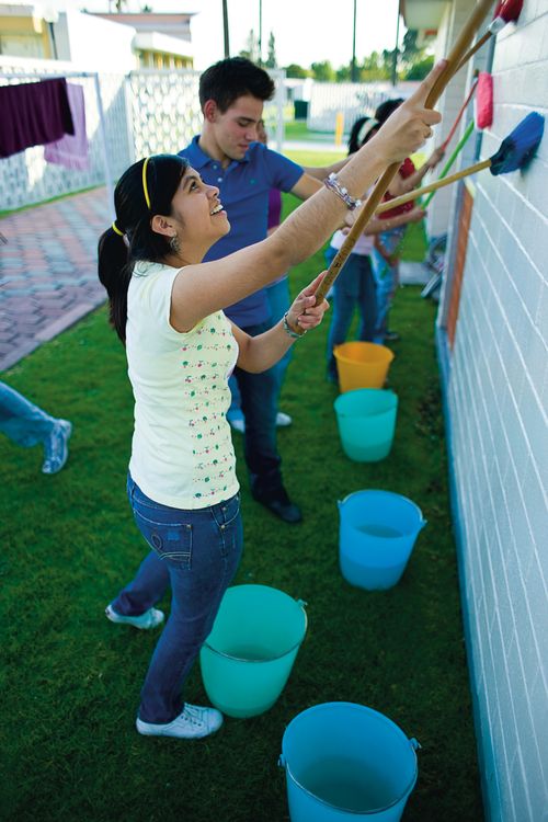 A group of youth wearing jeans and T-shirts work together to clean white walls outside with large brushes and buckets of water.