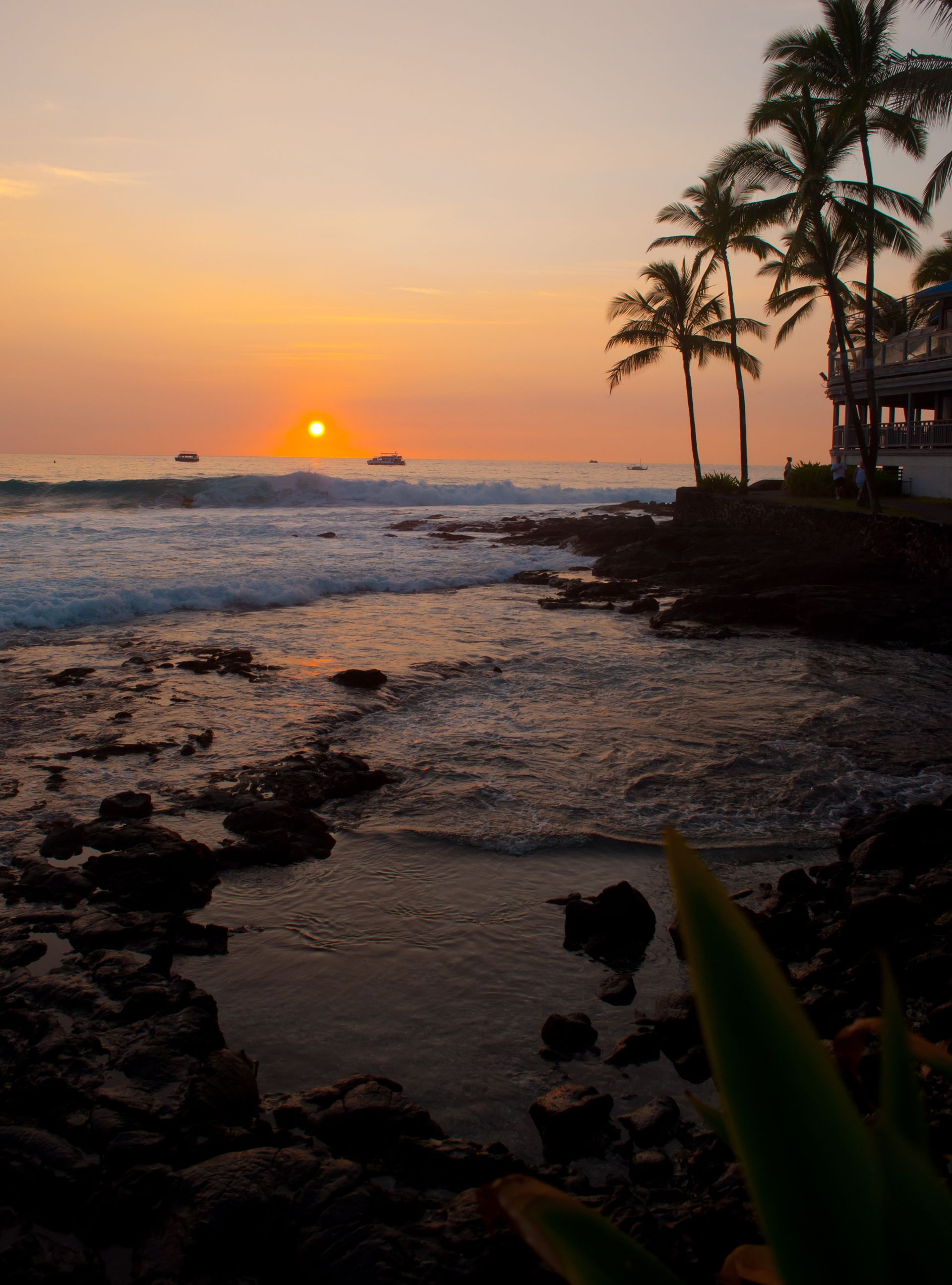 The sun sets over the ocean, with a view of the shore and palm trees.