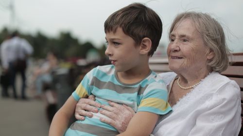 An elderly woman sits outside on a wooden bench with her young grandson.