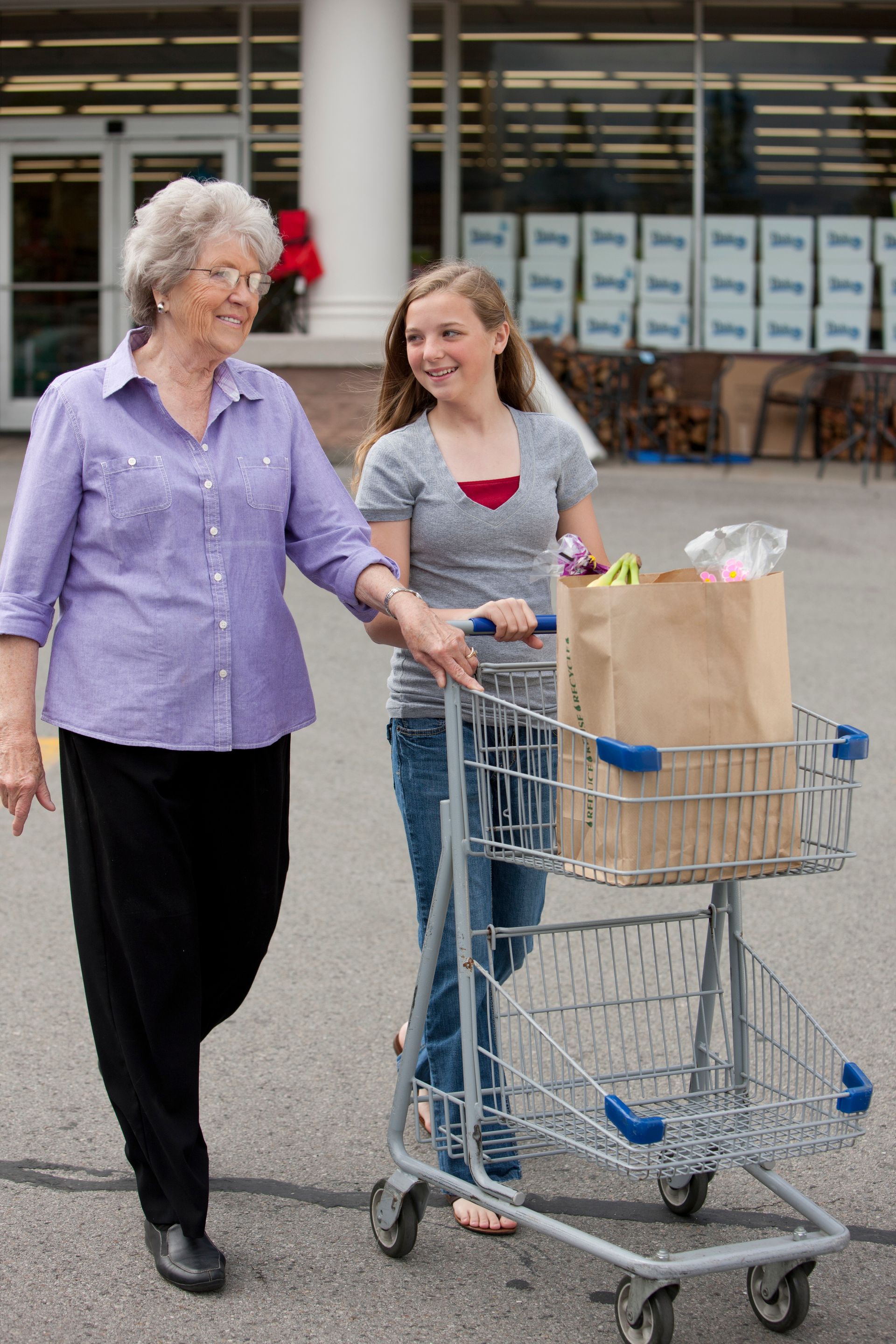 A young woman helps push a cart for an elderly woman with her groceries.  