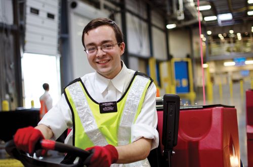 A young Church-service missionary in a yellow vest and red gloves drives a trolley in a warehouse.