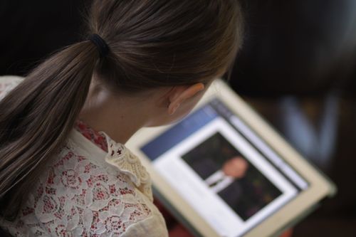 A young girl with long brown hair and a white lace shirt watches general conference on a tablet.