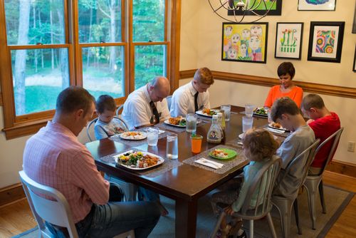 A father, mother, three sons, and a daughter sitting at a table and praying with two elder missionaries before eating their meal.