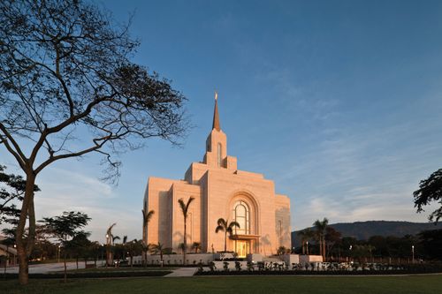 The entire San Salvador El Salvador Temple in the evening, including a view of the windows lit from inside, the grounds, and the trees surrounding the temple.