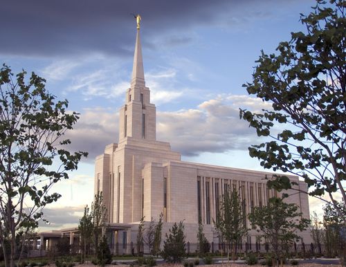 A side view of the Oquirrh Mountain Utah Temple, with small green trees in the foreground and other trees growing on the grounds near the temple.