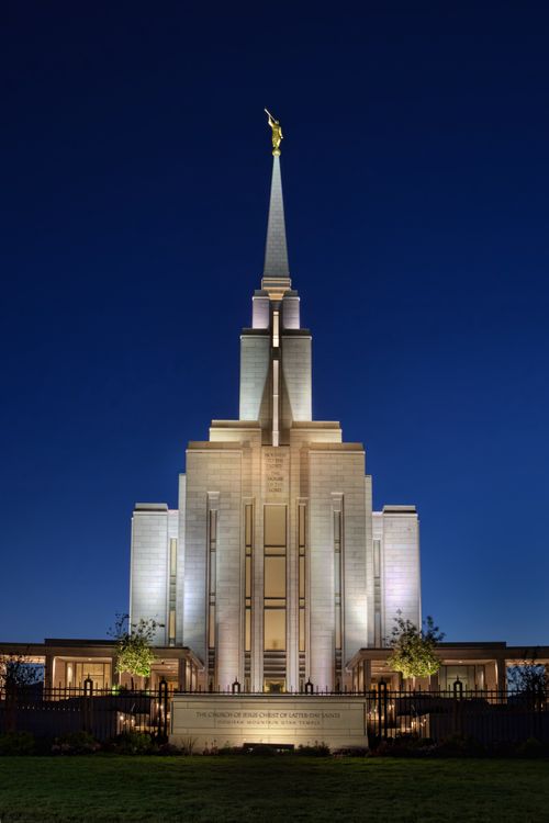 The front of the Oquirrh Mountain Utah Temple, with the lights on at night and a deep blue sky in the background.