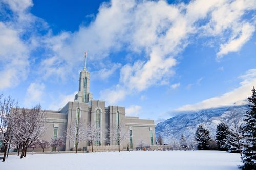 A view of the Mount Timpanogos Utah Temple and grounds covered in snow on a winter day, with fluffy white clouds overhead.