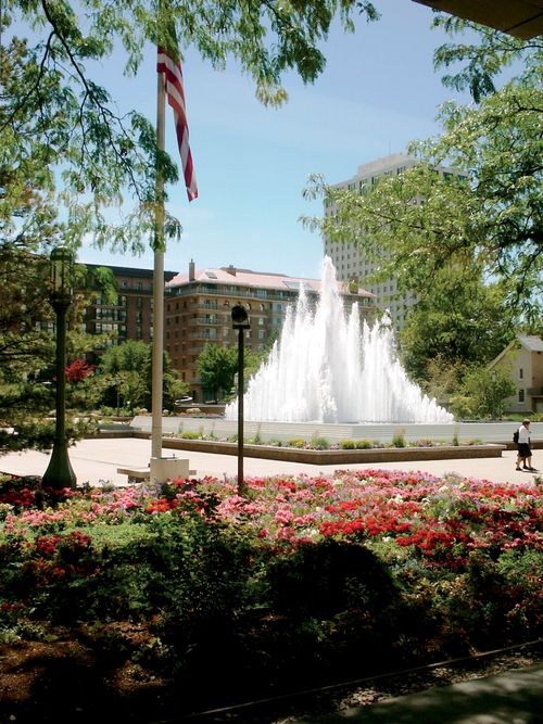 One of the fountains on Temple Square shooting water high into the air on a summer’s day, with a U.S. flag seen in the foreground.