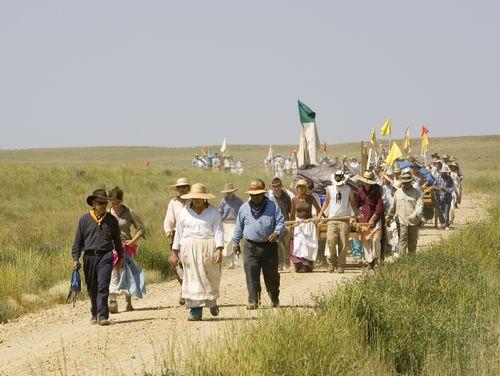 A large group of men and women, all dressed as pioneers in traditional clothing and hats, push handcarts with flags down a hill amid grassy plains.