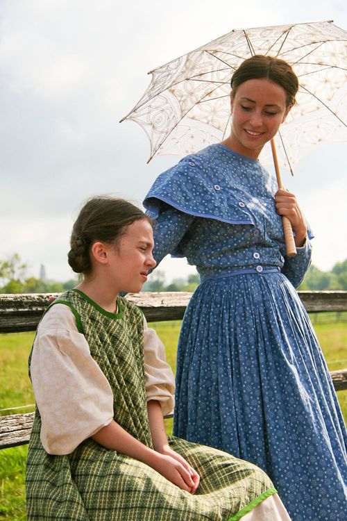 An adult actress from the Nauvoo Pageant in a blue dress, standing and holding a white umbrella while looking at a child actress who is sitting down.