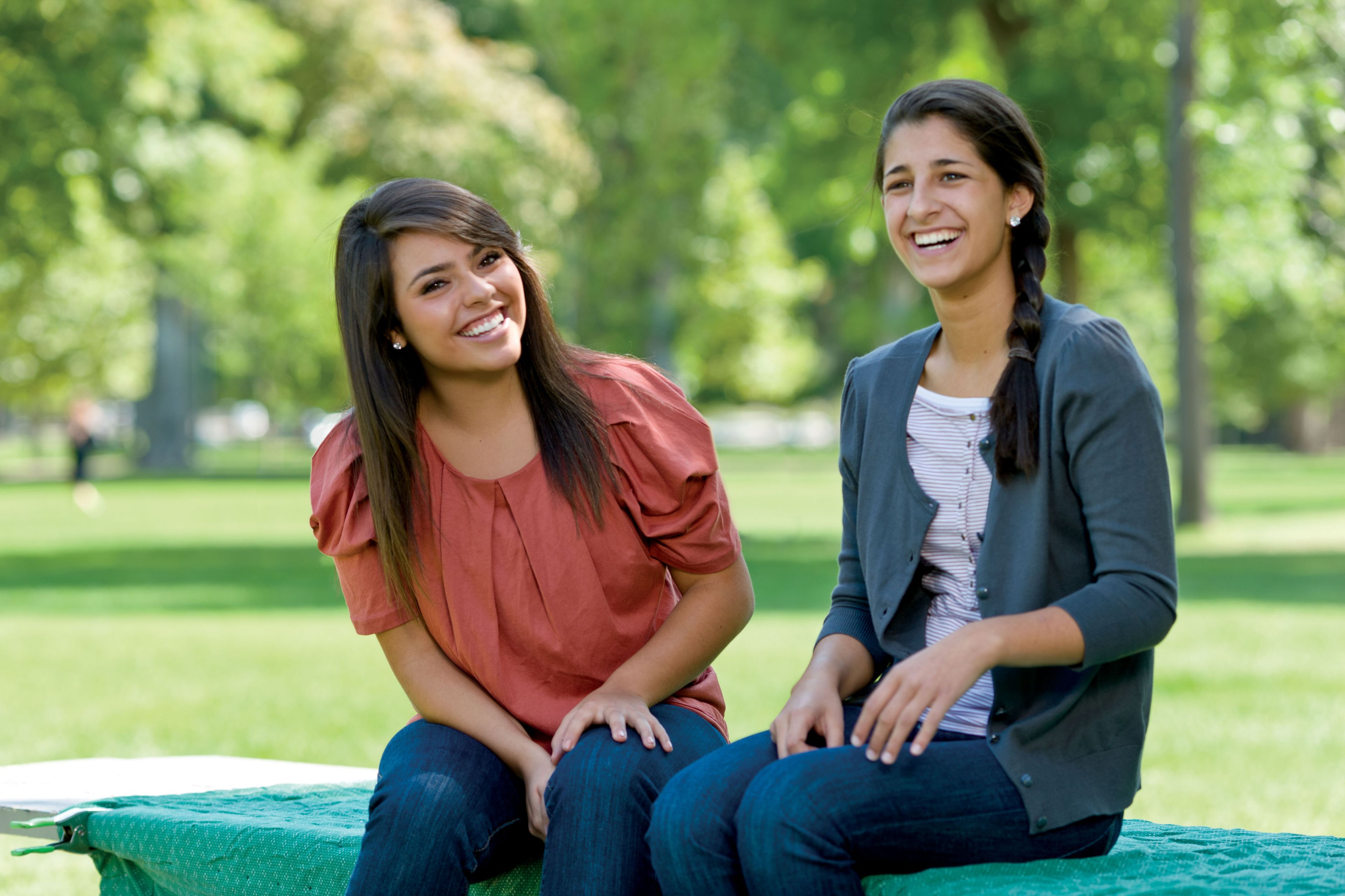 Two young women sitting together in a park.  They are laughing.