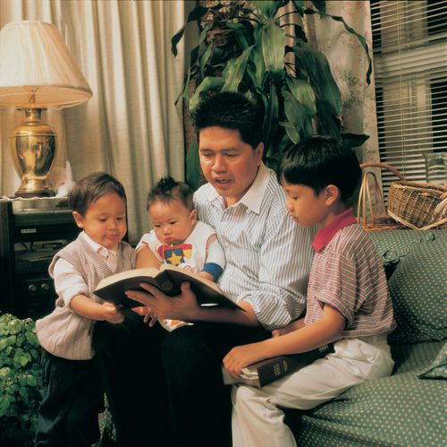 A father sits on a couch with his three young children around him and reads them a book.