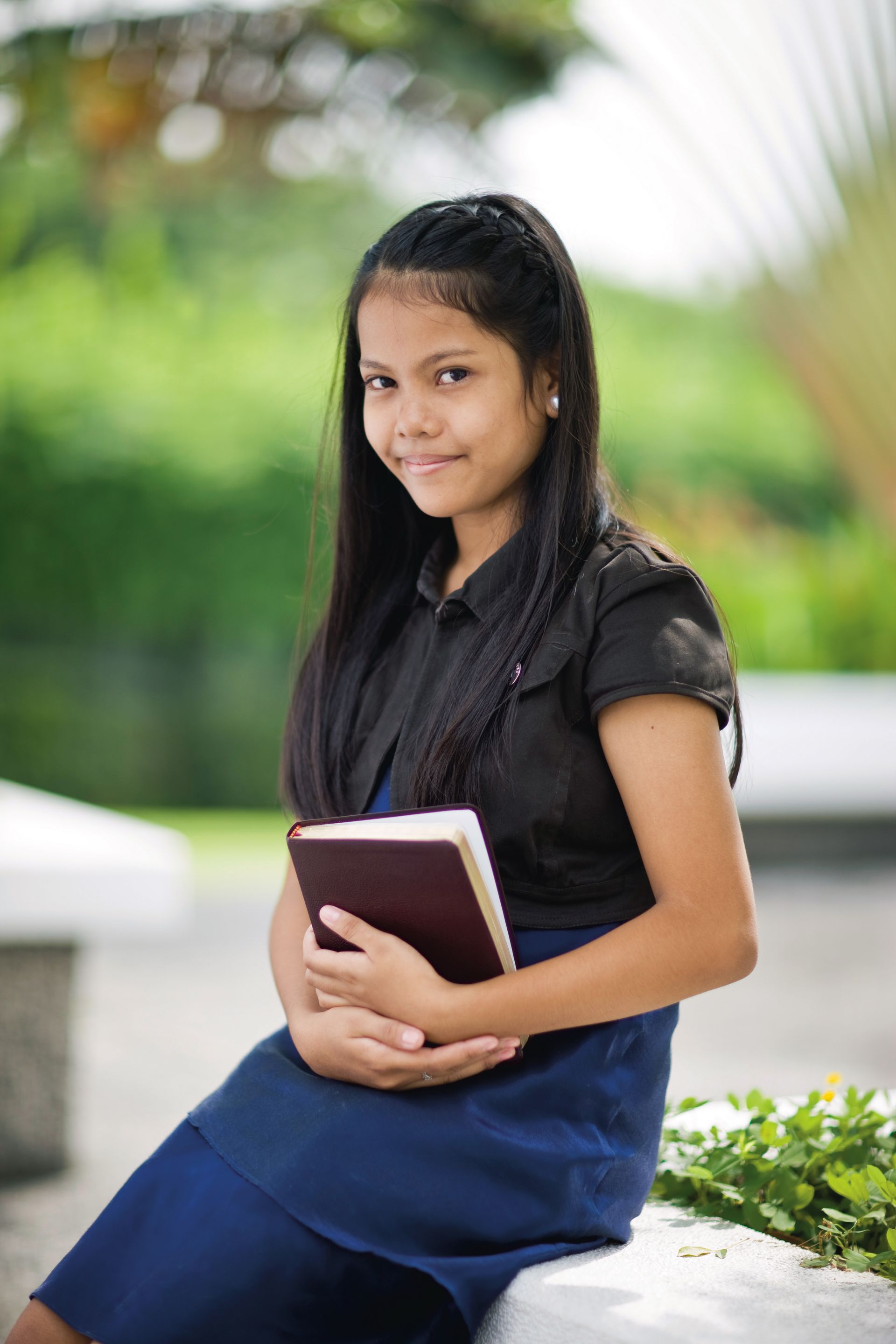 A young woman sits on temple grounds in the Philippines and smiles while holding her scriptures.