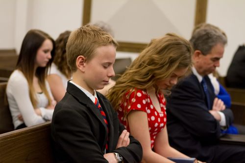 A family bow their heads and fold their arms while sitting in a pew at church and listening to a prayer.