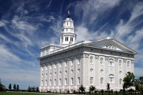 The back and side of the Nauvoo Illinois Temple on a bright day, with a blue sky and thin white clouds overhead.