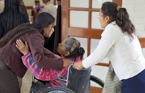 A woman pushing an elderly woman in a wheelchair.  Another woman is embracing the woman in the wheelchair.