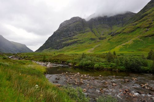 Fog lying low on mountains, with a stream running through a green, grassy valley in Scotland.