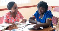 Various women sit around a table. On the table is a manual. It appears to be a manual helping them to learn English. The people read from the manual and share together. This is in Ghana, Africa.