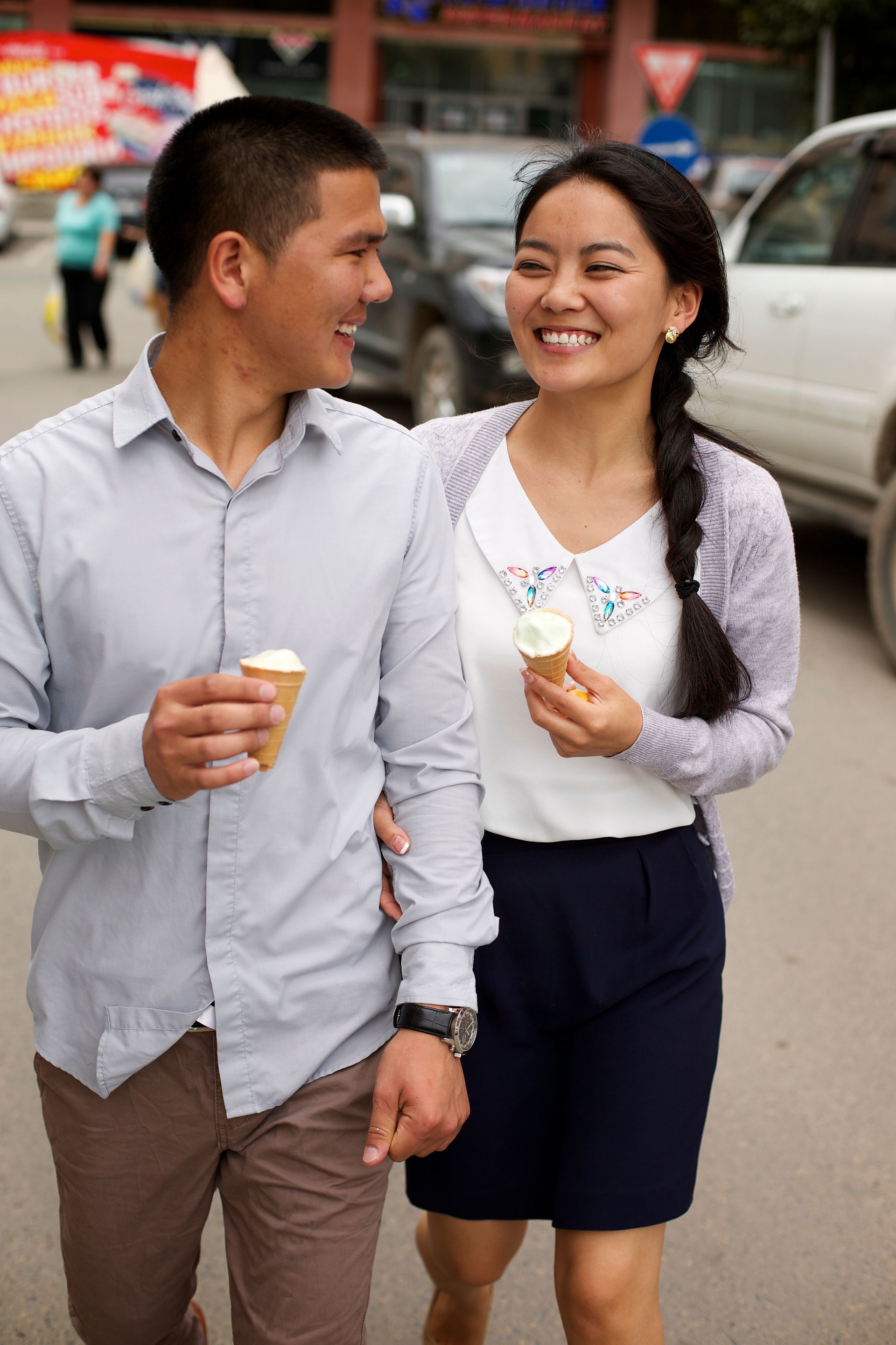 A young man and a young woman eat ice cream while walking down a street together.