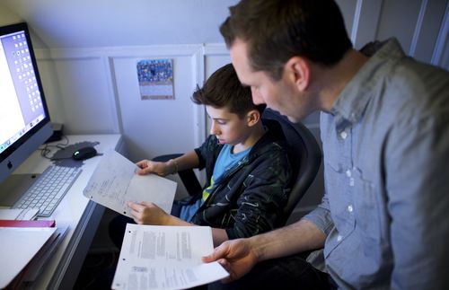 A father helps his son with his homework. A daughter also works on her own computer.
