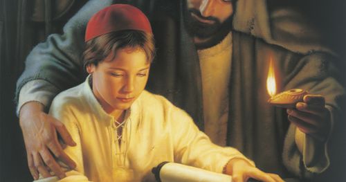 Jesus Christ depicted as a young boy in the carpenter shop of Joseph. Joseph is standing behind the young Christ. Christ is reading the scriptures from a scroll resting on a table. There are wood shavings on the table.