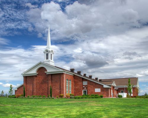 A green lawn surrounding a red-brick chapel with a white steeple and a blue sky with clouds overhead in Forest Grove.