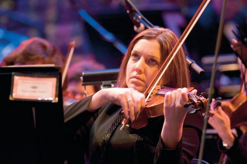 A female violinist sits in a chair and plays the violin while looking up at the conductor.