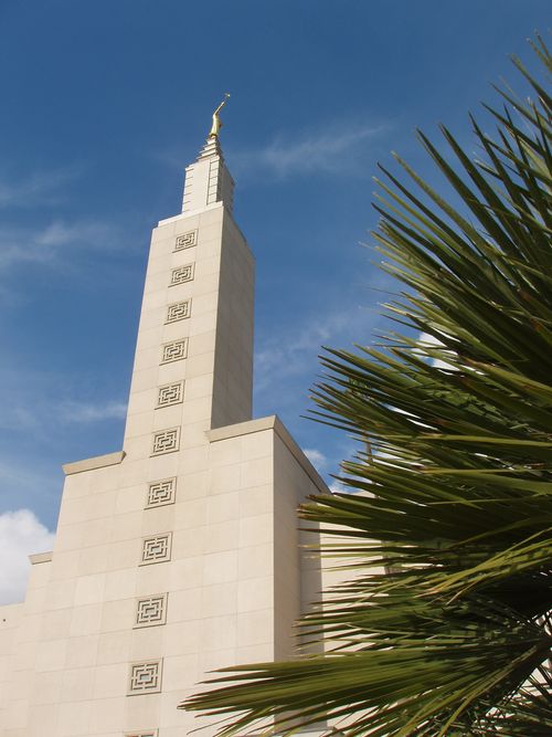 The spire on the Los Angeles California Temple, seen behind a green bush on the temple grounds, with a blue sky overhead.