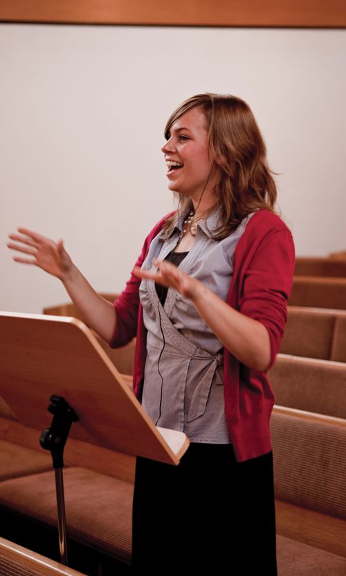 A young woman singing and standing behind a music stand while conducting a choir in song.