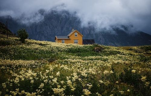 A house on a mountainside. there are flowers on the hill around the house. You can see the mountains in the background and clouds