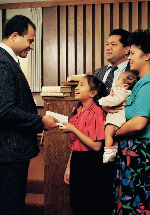 A young Polynesian girl handing her tithing to the bishop with her family standing behind her.