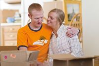 A young couple sitting next to each other in an apartment or house.  There are boxes around them to indicate they just moved in.  They are smiling and hugging each other.