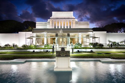 The fountain in front of the entrance to the Laie Hawaii Temple, at night, with the temple lit from inside and outside.