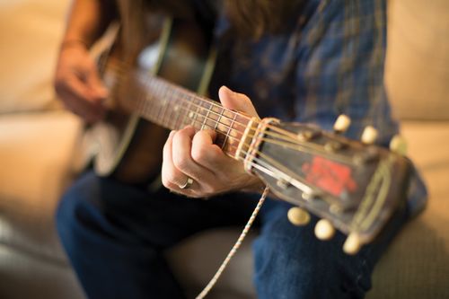 The hands of a young man are seen strumming a guitar while he sits on a couch.