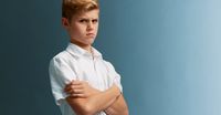 A young boy looking toward the camera. He is angry and upset. He has his arms folded, closing himself off. He is wearing a short sleeved white button down shirt.