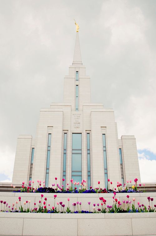 A front view of the Oquirrh Mountain Utah Temple on a spring day, with colored flowers in the front and a dull gray sky above.