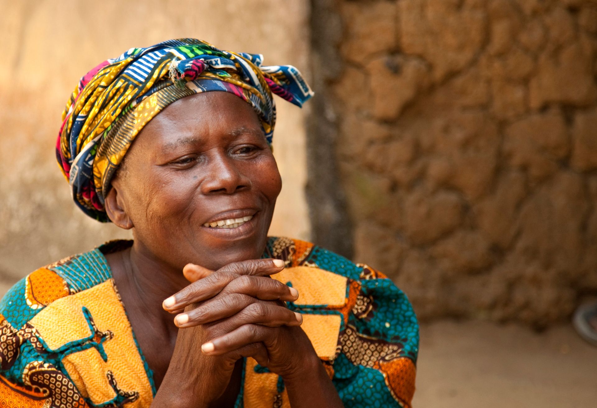 An African woman in a colorful head wrap and shirt clasping her hands together.