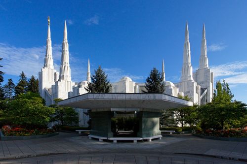 A side view of the Portland Oregon Temple showing all of the spires against a deep blue sky, surrounded by green trees.