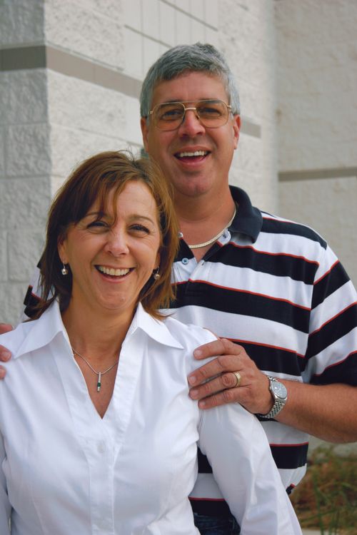 A portrait of a man in a black and white striped shirt standing behind his wife in a white shirt as they smile together.