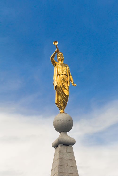 A view of the front of the angel Moroni statue standing on top of the spire of the Salt Lake Temple, with thin white clouds seen in the background.